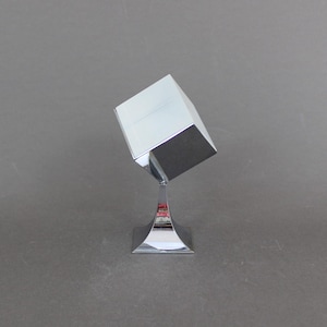 May include: A silver metal cube shaped box on a silver metal stand. The box is tilted at an angle.