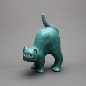 May include: A teal ceramic cat figurine in a standing, arched-back pose. The cat has a smooth, glossy finish, pointed ears, and wide, circular eyes with dark pupils. The tail is held upright, and the four paws are planted firmly on the surface.
