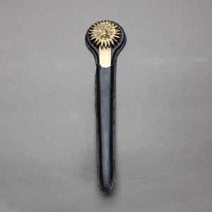 Könnte beinhalten: Ein schwarzes Lederwerkzeug mit einem goldfarbenen Sonnenmuster oben. Das Werkzeug hat einen langen, schlanken Griff und ein dekoratives Sonnenmuster. Das Werkzeug scheint ein Lederbearbeitungswerkzeug zu sein.