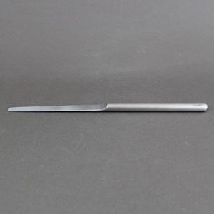May include: A slender, silver-toned metal letter opener with a smooth, cylindrical handle and a flat, tapered blade. The object features a minimalist design with a matte finish, extending horizontally across the frame with a rounded tip.