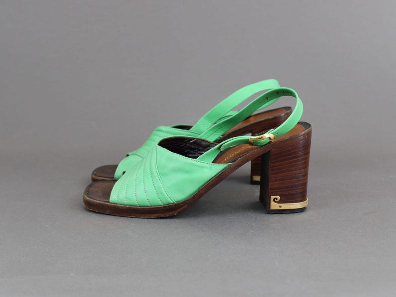 May include: A pair of green leather heeled sandals with a wooden platform and a gold buckle. The sandals have a criss-cross strap design.