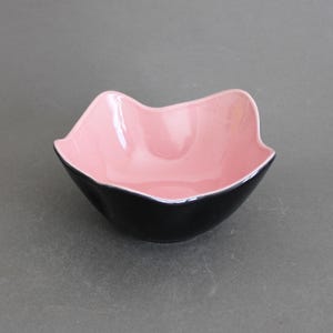 Puede incluir: Un cuenco pequeño y decorativo con una forma única. El interior es de color rosa brillante, mientras que el exterior es negro. Una fina línea plateada acentúa el borde. El cuenco está sobre una superficie gris.