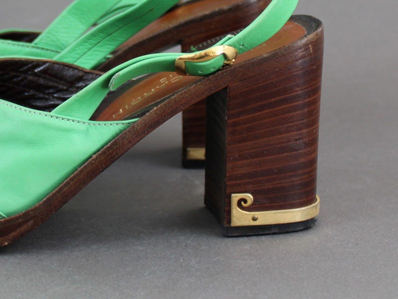 May include: Green leather heeled sandals with a wooden platform and a gold buckle. The sandals have a slingback strap and a closed toe.
