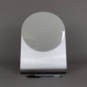 May include: A silver-coloured, freestanding vanity mirror with a round mirror surface.