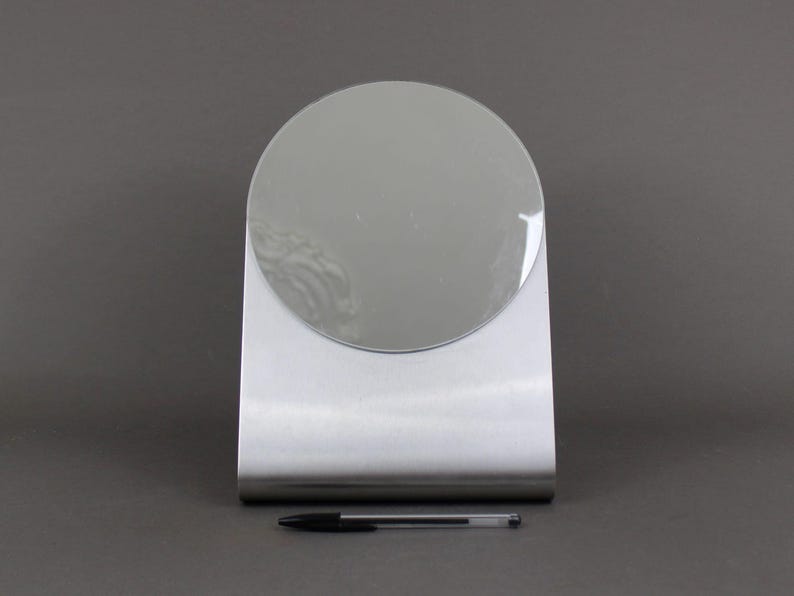 May include: A silver-coloured, freestanding vanity mirror with a round mirror surface.