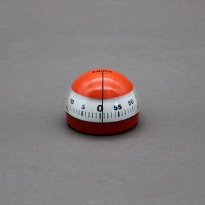 Peut inclure: Un minuteur de cuisine Krups vintage. Le minuteur a une base rouge, une section blanche avec des chiffres noirs et un dessus orange. Il compte jusqu'à 55 minutes. Le mot "KRUPS" est imprimé sur le dessus.