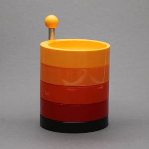 May include: A cylindrical container featuring a stack of five distinct color-blocked rings in shades of yellow, orange, red, dark red, and black. A small yellow sphere sits atop a silver metal post extending from the top yellow ring.