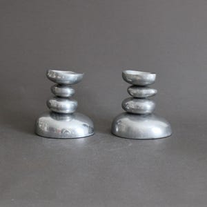 Peut inclure: Une paire de bougeoirs argentés, chacun composé d'une pile verticale de quatre formes lisses, arrondies et semblables à des galets. La base de chaque bougeoir est l'élément le plus large, surmonté de trois formes arrondies plus petites.