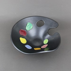 May include: A black ceramic artist's palette with a thumb hole and a small well. The palette has colourful paint wells in shades of green, red, yellow, blue, and orange.