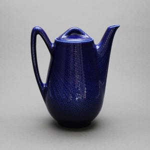 Peut inclure: Une cafetière en céramique bleu cobalt avec un couvercle et une poignée incurvée. Le pot présente un motif en chevrons texturé. Le bec est incurvé et la poignée est arquée. Le pot est sur fond gris.