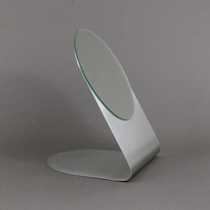 May include: A silver-coloured, freestanding vanity mirror with a round, slightly angled glass surface.