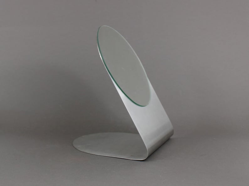 May include: A silver-coloured, freestanding vanity mirror with a round, slightly angled glass surface.