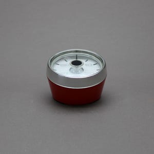May include: A circular analog gauge featuring a white face with black numerical markings and a central needle. The gauge is encased in a polished silver-tone metal bezel and has a solid, deep red cylindrical base, presenting a compact design.