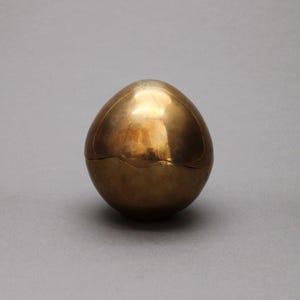 Könnte beinhalten: Ein goldfarbener, eiförmiger Behälter mit Scharnierdeckel. Der Behälter hat eine glatte, reflektierende Oberfläche und einen warmen, metallischen Farbton. Das Ei liegt auf einem neutralen grauen Hintergrund.