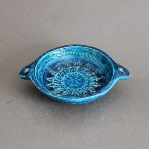Op de afbeelding: Een kleine, ronde keramische kom met een levendige turquoise glazuur. De kom heeft twee kleine handvatten en een gedetailleerd, verhoogd bloemmotief in het midden. De binnen- en buitenkant zijn diep, rijk blauw.