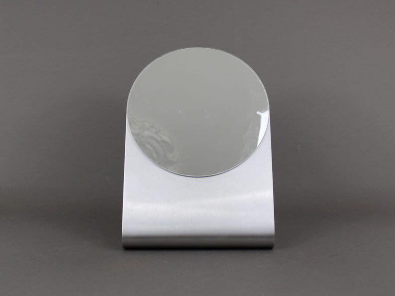 May include: A silver-coloured, freestanding tabletop mirror with a round, reflective surface.