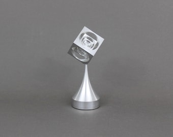 Könnte beinhalten: Eine moderne, abstrakte Skulptur aus silberfarbenem Metall. Die Skulptur zeigt einen Würfel mit verschachtelten kreisförmigen Aussparungen, der auf einem konischen Stiel und einer kreisförmigen Basis balanciert. Das Kunstwerk wird vor einem grauen Hintergrund präsentiert.
