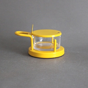 May include: A yellow condiment server with a clear glass container. The server has a round base, a lid, and a handle. The handle is attached to the side of the server. The server is made of metal and glass.