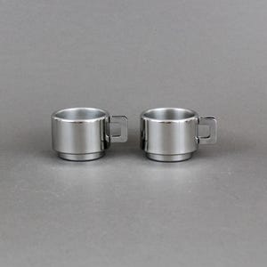 Pode incluir: Duas pequenas canecas de aço inoxidável. Cada caneca tem um corpo cilíndrico e uma pequena alça quadrada. As canecas são empilhadas, com uma base ligeiramente menor. O fundo é cinza liso. Adequado para café expresso.