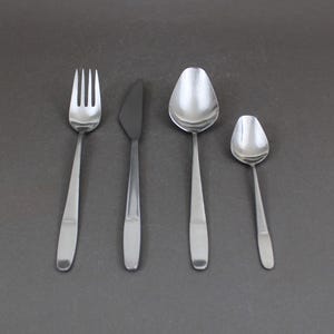 May include: A collection of four stainless steel cutlery items: a fork, a knife, a soup spoon, and a teaspoon. The fork has four prongs, the knife has a rounded end, and the spoons have oval bowls. The items are on a dark grey surface.
