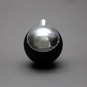 May include: A spherical black container with a polished chrome rotating lid. The object features a smooth matte black finish on the lower half and a reflective metallic dome top that pivots open, resting on a small circular base.