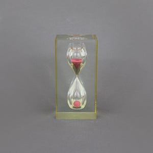 May include: A clear hourglass with a pink sand timer inside. The hourglass is in a clear plastic frame.