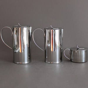 Può includere: Un set di tre pezzi da portata in acciaio inossidabile, tra cui due brocche con manici curvi e una piccola lattiera o zuccheriera. Gli articoli sono altamente riflettenti, con una finitura lucida, e poggiano su una superficie grigia.