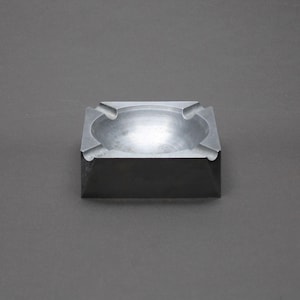 Può includere: Un posacenere quadrato grigio con quattro rientranze curve per appoggiare le sigarette. Il posacenere ha una base leggermente smussata ed è realizzato in un materiale solido, possibilmente metallico. Il posacenere è centrato su uno sfondo grigio.