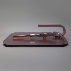 May include: A pink glass cheese board with a silver cheese knife. The knife is resting on the board and has a handle made of pink glass.