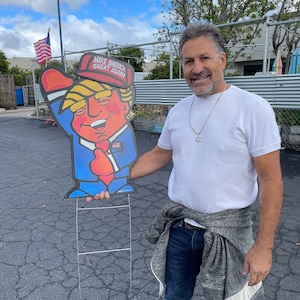 May include: A person holding a cardboard cutout of a cartoon figure wearing a red hat with the text "Make America Great Again". The figure is wearing a blue suit and red tie. The person is wearing a white t-shirt, blue jeans, and a grey sweater tied around their waist.