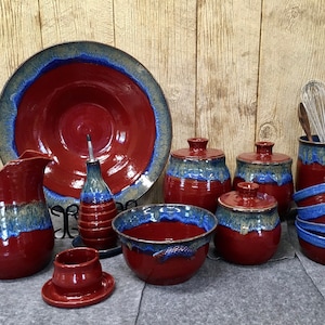 May include: A set of handmade pottery kitchenware in red and blue glaze. The set includes a large serving bowl, a pitcher, a small bowl, a butter dish, a small oil bottle, three canisters with lids, and two bowls.