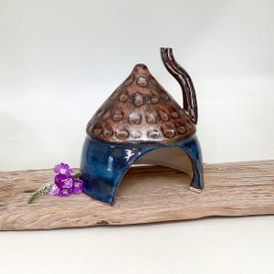 May include: A ceramic dome-shaped structure with a glossy blue base and a textured, metallic bronze-toned top featuring a curved handle. The object has an arched opening at the front and is accompanied by a small cluster of purple flowers.