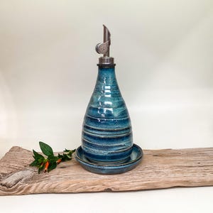 May include: A teardrop-shaped ceramic oil cruet with a glossy, variegated blue glaze featuring horizontal ridges. The bottle is topped with a stainless steel pourer spout and rests on a matching circular ceramic saucer.