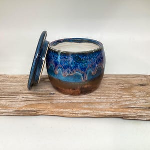 May include: A handmade ceramic jar with a lid, featuring a glossy, drippy glaze in shades of blue, turquoise, and brown. The jar is round with a wide opening, and the lid is slightly off to the side. The jar sits on a piece of weathered wood.