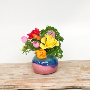 May include: A small, colorful ceramic vase with a pink, blue, and green glaze. The vase is filled with a bouquet of pink, yellow, and orange flowers.