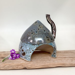 May include: A ceramic dome-shaped structure with a glossy finish, featuring a mottled blue and grey glaze with dark brown speckles. The piece has a curved, upright handle on the right side and an arched opening at the base for entry or exit.