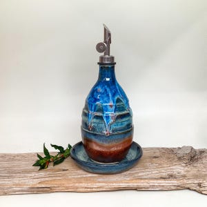 May include: A ceramic oil cruet with a metallic pour spout, featuring a glossy glaze that transitions from deep blue at the top to a warm reddish-brown at the base. The bottle sits on a matching circular ceramic saucer.