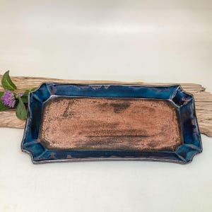 May include: A rectangular ceramic tray with a dark blue rim and a copper-colored interior. The tray has a decorative, scalloped edge. A sprig of purple flowers and green leaves are on the left side of the tray.