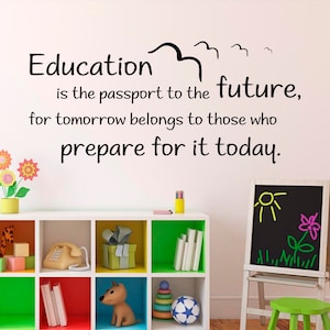 May include: A white wall with a black motivational quote that reads "Education is the passport to the future, for tomorrow belongs to those who prepare for it today." The quote is decorated with a black bird illustration. A white bookshelf with colorful compartments is in the foreground. The bookshelf is filled with toys and books. A black chalkboard easel with a yellow sun and pink flower drawing is on the right side of the image.