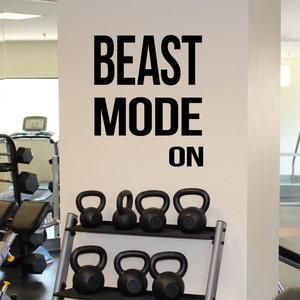 Puede incluir: Una escena de gimnasio con la frase "BEAST MODE ON" en letras negras grandes en una pared. Debajo, una estanter&iacute;a met&aacute;lica contiene varias pesas rusas negras de diferentes tama&ntilde;os. Otros equipos de gimnasio, como pesas y una cinta de correr, son visibles.
