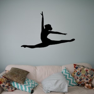 Op de afbeelding: Een zwart silhouet van een danseres in een splits, met één been gestrekt en het andere been gebogen bij de knie. De armen van de danseres zijn boven haar hoofd geheven. Het silhouet bevindt zich op een lichtblauwe muur, boven een witte bank met kussens.