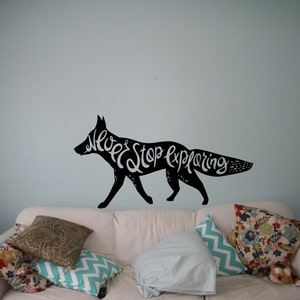 Può includere: Un adesivo murale nero a forma di volpe con le parole "Never Stop Exploring" in corsivo bianco. L'adesivo è su una parete azzurra sopra un divano bianco con cuscini decorativi.