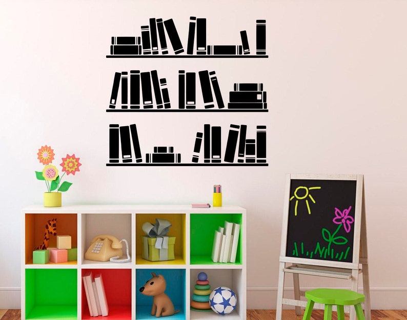 May include: Black and white silhouette of bookshelves with books on each shelf. The shelves are on a white wall. There is a small wooden easel with a black chalkboard in front of the shelves. The chalkboard has a yellow sun and a pink flower drawn on it.