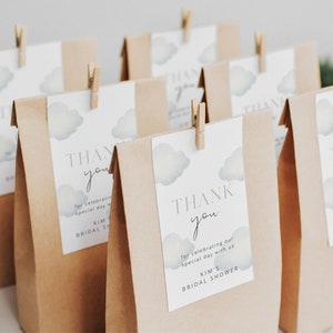 May include: Brown paper bags with thank you tags attached with clothespins. The tags feature a watercolor design of clouds and the text "Thank you for celebrating our special day with us. Kim's Bridal Shower."
