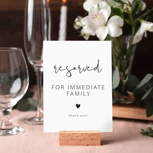 Könnte beinhalten: Eine weiße Karte mit schwarzem Text, der "reserviert für die unmittelbare Familie" lautet, mit einem kleinen schwarzen Herzen und den Worten "Danke!" auf einem Holzständer. Die Karte steht auf einem Tisch mit zwei Gläsern, Kerzen und Blumen.