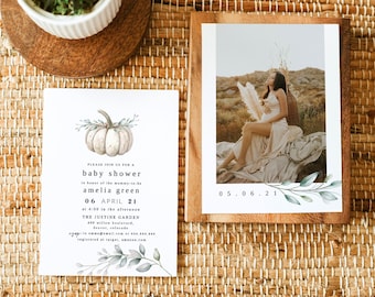 May include: A baby shower invitation with a watercolor pumpkin design and text. The invitation is next to a framed photograph of a pregnant person. A small potted plant sits on a woven mat.