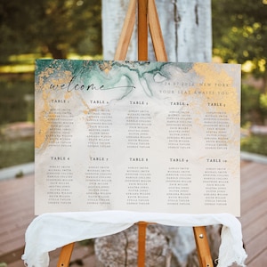 May include: A wooden easel displays a wedding seating chart. The sign features a watercolor design with gold and teal accents, the word "welcome", and guest names listed under table numbers. The date and location are also displayed.