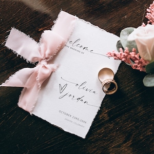 May include: A wedding invitation with the text "welcome to the wedding of Olivia Jordan" and a pink ribbon. Two gold wedding rings are next to the invitation. A boutonniere with a pink rose and greenery is also present.