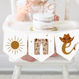 May include: A banner with three white pennants, each featuring a different design in brown. The designs include a sun, pants, and a cowboy hat with the letter "E" and a gun. The banner is part of a first birthday celebration.