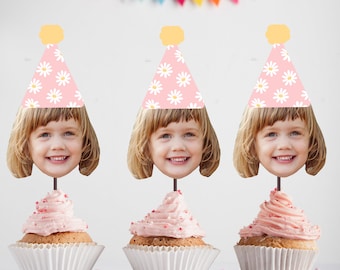 May include: Three pink cupcakes with pink frosting and sprinkles topped with a cupcake topper featuring a child's face wearing a pink and white daisy party hat.